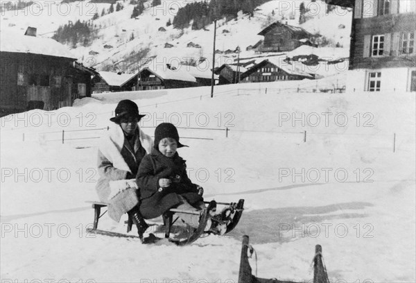 Indian ayah holidaying with family. An Indian ayah (nursemaid) called Nannie sits on a sledge with her charge, Owen, at a snowy holiday resort. Nannie worked for a British family (the Lawrences) in India, later moving with them to England where she lived for the rest of her life. Location unknown, circa 1919.