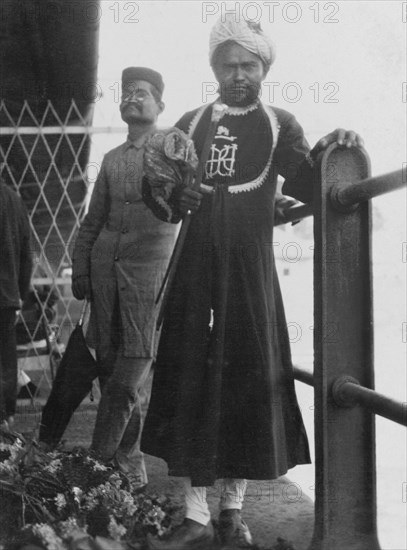 Indian man in ceremonial dress. Portrait of a turbaned Indian man in ceremonial dress. He wears an embroidered 'achkan' (long jacket) with an emblem on the breast, over 'churidar' (fitted trousers), and holds up a short staff. India, circa 1910. India, Southern Asia, Asia.