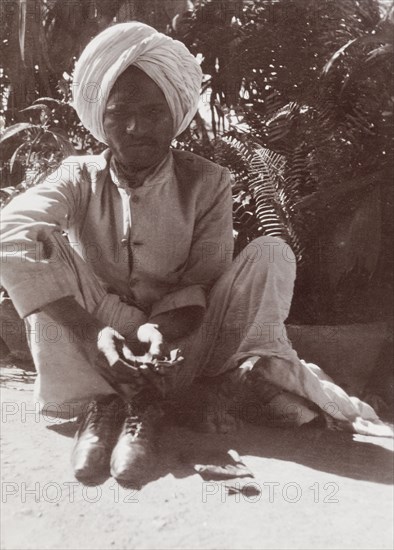 Indian man in Karachi. An Indian man squats on a street in Karachi, barefoot with his shoes positioned in front of him. Karachi, Sind, India (Sindh, Pakistan), circa 1910. Karachi, Sindh, Pakistan, Southern Asia, Asia.