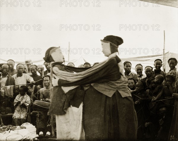 A masked performance. An amused audience looks on as two masked figures perform at an outdoor festival. An original caption suggests they are wrestling, but this may instead be a variation on Korean mask dancing. Probably China, circa 1910. China, People's Republic of, Eastern Asia, Asia.