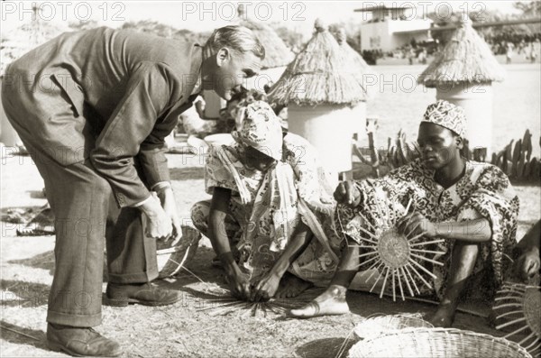 A basket-weaving demonstration. A European man visiting Kaduna Children's Village with Queen Elizabeth II, stoops down to watch a group of young men demonstrate traditional basket-weaving skills outdoors. Kaduna, Nigeria, February 1956. Kaduna, Kaduna, Nigeria, Western Africa, Africa.