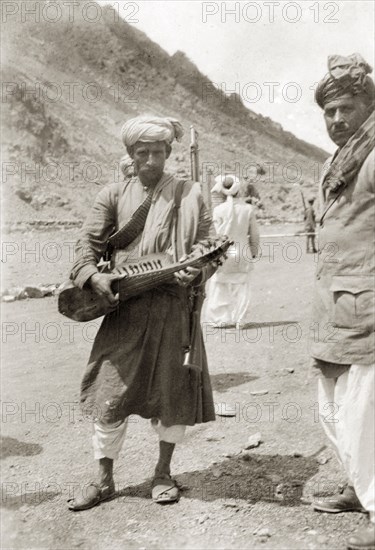 Playing a sarod at the border. An Indian man, armed with a rifle and wearing a bandolier, plucks a sarod (Indian stringed instrument) at a military checkpoint on the Afghanistan-India (Pakistan) border. India (Pakistan), circa 1930. Pakistan, Southern Asia, Asia.