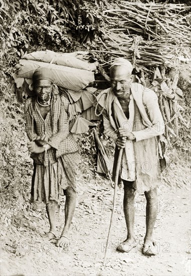 Indian couple on a rural road. An Indian couple walk along a road, carrying cumbersome bundles of leaves and firewood on their backs. India, circa 1915. India, Southern Asia, Asia.