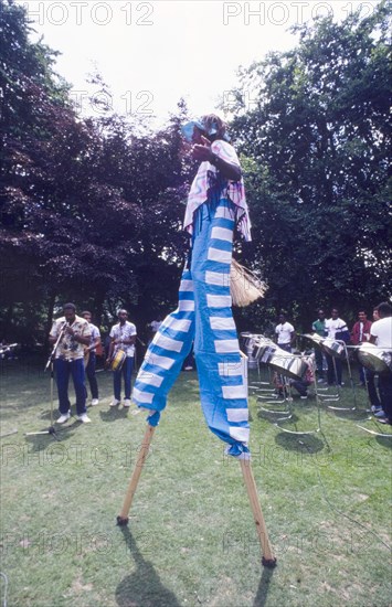 Stilt walker at the Caribbean Music Village. A stilt walker dressed in a blue and white striped costume performs at the Caribbean Music Village, a cultural festival held at the Commonwealth Institute. London, England, 7-26 July 1986. London, London, City of, England (United Kingdom), Western Europe, Europe .