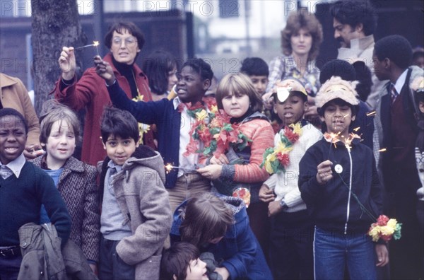 Children light sparklers at an outdoor carnival. A group of schoolchildren wearing masks and floral garlands light sparklers as part of festivities for a Commonwealth Institute carnival, possibly a celebration for Diwali, the Hindu festival of light. London, England, circa 1985. London, London, City of, England (United Kingdom), Western Europe, Europe .