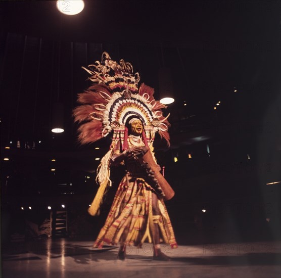 Papua New Guinean performance. A male dancer dressed in a ceremonial costume from Papua New Guinea plays a hand drum as he performs on stage during a cultural festival at the Commonwealth Institute. His face is painted yellow and he wears a large, elaborately feathered headdress. London, England, circa 1985. London, London, City of, England (United Kingdom), Western Europe, Europe .