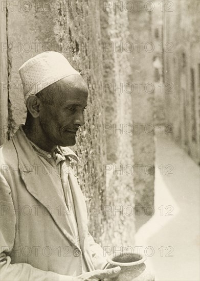 Profile shot of a Muslim man. Profile shot of a Muslim man wearing an embroidered kufi (cap). He leans against a stone wall on a narrow street, holding up a clay pot. Probably Lamu, Kenya, circa 1947. Lamu, Coast, Kenya, Eastern Africa, Africa.