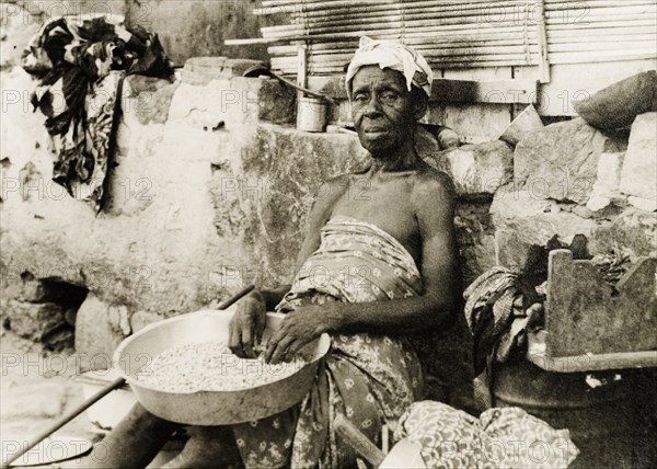 An elderly street vendor in Accra. An elderly woman sits against a roadside wall, selling nuts or grains from a bowl. Accra, Gold Coast (Ghana), circa 1950. Accra, East (Ghana), Ghana, Western Africa, Africa.