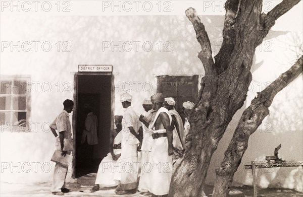 Queueing to see the District Officer. A group of Somali men queue outside an office building for an appointment with the District Officer. Wajir, Norther Frontier District (North Eastern Province), Kenya, circa 1935., North East (Kenya), Kenya, Eastern Africa, Africa.