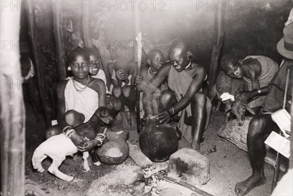 Evening meal in a Kikuyu hut. A Kikuyu woman serves an evening meal to her family, who are gathered around a fire inside their hut with a number of goat kids. South Nyeri, Kenya, 1936. Nyeri, Central (Kenya), Kenya, Eastern Africa, Africa.