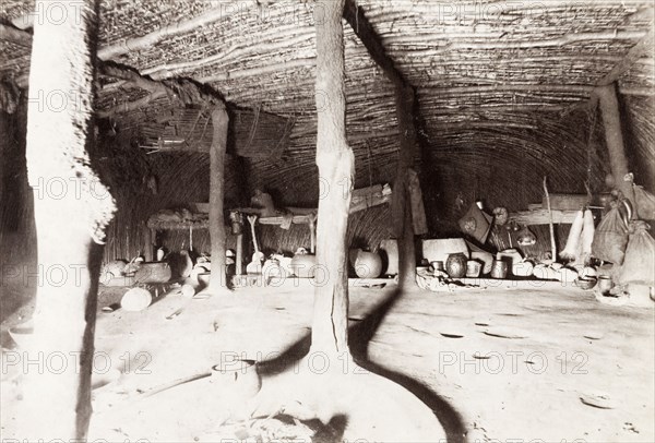 Interior of a traditional Zulu dwelling. The spacious interior of a traditional Zulu dwelling. A framework of beams supports the dome-shaped roof, which is thatched with thick grass mats. A variety of cooking pots and gourds sit against the far wall. Probably Natal (KwaZulu-Natal), South Africa, circa 1920. South Africa, Southern Africa, Africa.