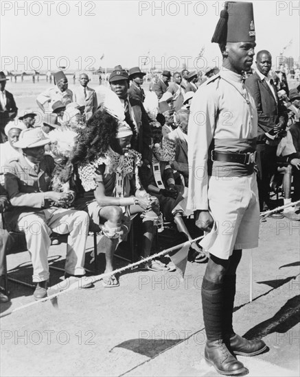 Awaiting the arrival of Princess Elizabeth. An askari (soldier) stands to attention in front of a spectator stand, where an African assembly awaits the arrival of Princess Elizabeth at Eastleigh Airport. Nairobi, Kenya, February 1952. Nairobi, Nairobi Area, Kenya, Eastern Africa, Africa.