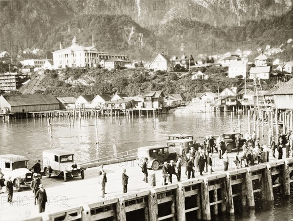 Juneau Pier, Alaska. Visitors enjoy the view of the Gastineau Channel from the wooden pier at the Alaskan capital city of Juneau, which can be seen nestled in the foothills of the mountains beyond. Juneau, Alaska, United States of America, circa 1931. Juneau, Alaska, United States of America, North America, North America .