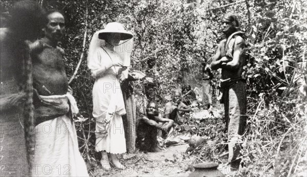 Ceylonian beaters on elephant drive. A European woman stands in a jungle clearing with a team of Ceylonian beaters, as they take a break from driving elephants into a kraal (enclosure). The lady wears a wide-brimmed hat draped with mosquito netting, and a dress with the outer skirt hiked up around her knees. Ceylon (Sri Lanka), 1912. Sri Lanka, Southern Asia, Asia.