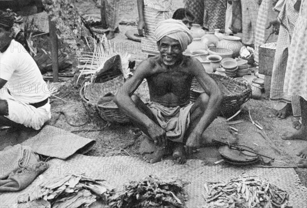 Street trader in Ceylon. A street trader sells a variety of dried foods at a busy outdoor market. Ceylon (Sri Lanka), circa 1935. Sri Lanka, Southern Asia, Asia.