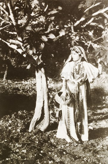 Portrait of a Palestinian woman. Portrait of a Palestinian woman posing beside a tree in a village near Ramallah. She wears an embroidered robe and headscarf, and holds a basket in one hand decorated with bangles and rings. Ramallah, British Mandate of Palestine (West Bank, Middle East), circa 1942. Ramallah, West Bank, West Bank (Palestine), Middle East, Asia.