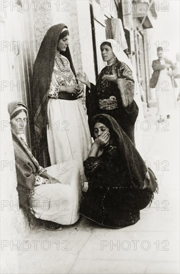 Four Arab women in Bethlehem. Four Arab women chat outside a doorway on a city street. All wear traditional Palestinian dress including embroidered 'thobs' and headscarves. Bethlehem, British Mandate of Palestine (West Bank, Middle East), circa 1938. Bethlehem, West Bank, West Bank (Palestine), Middle East, Asia.