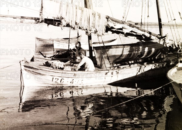 Fishing boat on the Suez Canal. Two fishermen sit aboard a stationary sail boat on the Suez Canal, beneath a row of fishing nets hung up over the boom. Suez, Egypt, circa 1930. Suez, Suez, Egypt, Northern Africa, Africa.