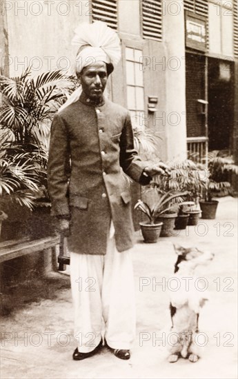 A 'Gentlemen's Gentleman'. Full-length portrait of an Indian valet named Abdul, employed by British businessman James Murray who describes him as "my Gentlemen's Gentleman". He poses playfully with the Murrays' pet dog George, wearing his house uniform and a fanned turban. Calcutta (Kolkata), India, circa 1935. Kolkata, West Bengal, India, Southern Asia, Asia.