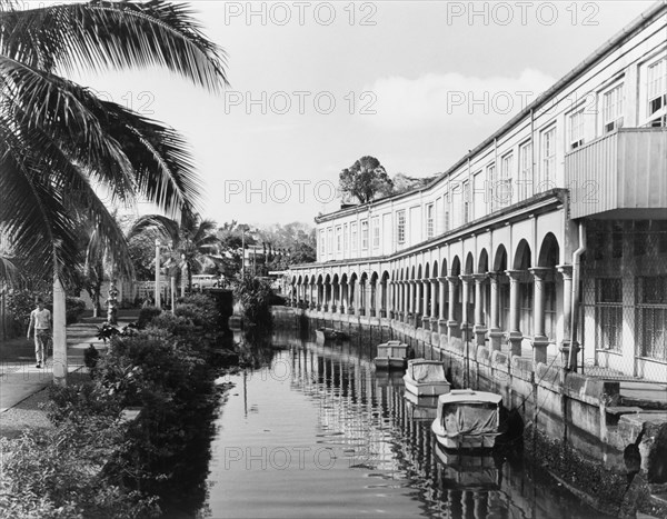 A waterway in Suva. An arcade borders a waterway in central Suva, where several small boats are moored. Suva, Fiji, 1965. Suva, Viti Levu, Fiji, Pacific Ocean, Oceania.