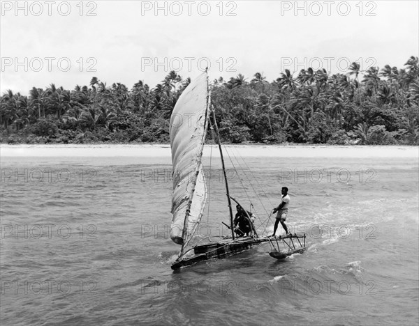 Manning a Fijian 'drua'. A 'drua' (Fijian catamaran) scuds across the waves away from a beach flanked by palm forest. Fiji, 1965. Fiji, Pacific Ocean, Oceania.