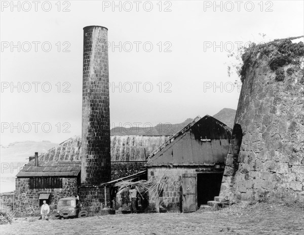 A sugar mill in Monserrat. A brick chimney and outbuildings form part of a sugar mill that was once the island's principal rum factory. Two men can be seen carrying harvested sugar cane into one of the buildings. Monserrat, 1965. Montserrat, Caribbean, North America .