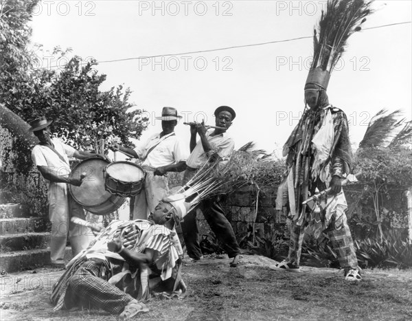 Carnival dancers on Nevis. Two men perform a carnival dance outdoors, accompanied by a musical band. Both dancers wear crowns adorned with peacock feathers and costumes made from ribbons and scarves. Nevis, 1965. St Kitts and Nevis, Caribbean, North America .