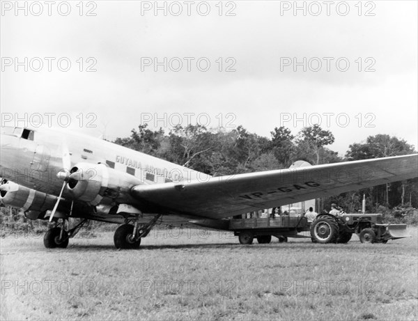 A Guyana Airways Corporation aeroplane . A Guyana Airways Corporation aeroplane sits in a grassy field as it is loaded with cargo from the back of a tractor. An original caption comments that the aircraft was "en route to the interior". British Guiana (Guyana), 1965., Guyana, South America, South America .