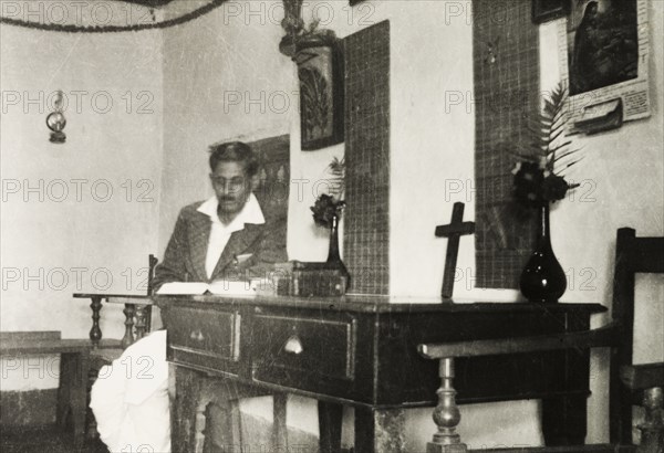 House prepared for a Christian service. The interior of a house on a coffee estate prepared for a Christian church service. A Methodist evangelist sits reading at the desk, which is set up as an altar containing a cross, bible and flowers. Mysore State (Karnataka), India, circa 1937., Karnataka, India, Southern Asia, Asia.