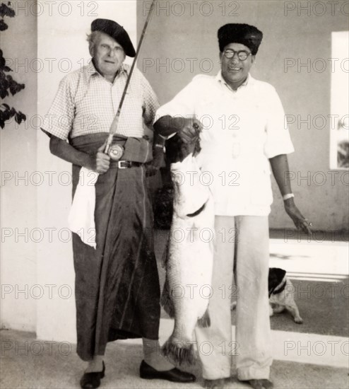 Prize fish from West Bengal. Frank Snaith poses proudly for the camera, dressed in a traditional Indian 'lungi' (wraparound skirt) and holding a fishing rod. Beside him, a turbaned Indian man holds up a huge fish, which Snaith caught in the Bay of Bengal. Digha, West Bengal, India, February 1953. Digha, West Bengal, India, Southern Asia, Asia.