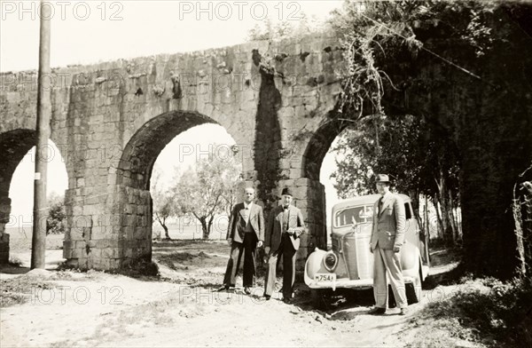 Visiting the aqueduct near Acre. Three men in suits stand beside a car beneath the arches of the old Roman aqueduct near Acre. Near Acre (Akko), British Mandate of Palestine (Northern Israel), circa 1940. Akko, North (Israel), Israel, Middle East, Asia.