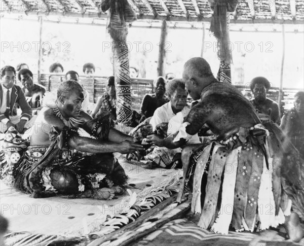 The culmination of a 'sevusevu' ceremony. A 'bilo' (half coconut shell) containing an infusion of water and powdered kava root (Piper methysticum) is passed between two men during a traditional 'sevusevu' welcoming ceremony. Watched by a seated audience, both men are naked from the waist up and wear ceremonial costume including face paint and skirts made from strips of fabric. Fiji, 1965. Fiji, Pacific Ocean, Oceania.