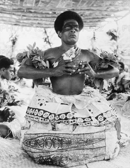 Preparing to drink a kava infusion. A Fijian man prepares to drink an infusion of water and powdered kava root (Piper methysticum) from a 'bilo' (half coconut shell) during a traditional 'sevusevu' welcoming ceremony. Naked from the waist up, he wears ceremonial costume including dramatic face paint and a tiered 'masi' (bark cloth) skirt. Fiji, 1965. Fiji, Pacific Ocean, Oceania.