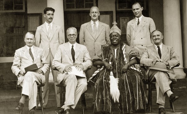 Western officials with a Nigerian chief. Western officials pose for a group portrait with a Nigerian chief, possibly Obafemi Awolowo, who is dressed in full royal regalia. Ife, Osun State, Nigeria, October 1942. Ife, Osun, Nigeria, Western Africa, Africa.