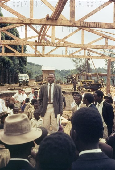 Mr Bitware speaks at Mafuga sawmill. Mr J. Bitwari addresses a crowd of Ugandan chiefs, villagers and schoolchildren during a forestry open day at Mafuga sawmill. Above him is a partly finished mill building comprising upright beams and roof trusses. Mafuga, Kigezi, South West Uganda, September 1963., West (Uganda), Uganda, Eastern Africa, Africa.