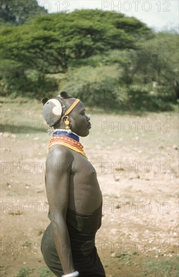 Portrait of a Suk porter. Profile portrait of a Suk (Pokot) porter working with a British forestry survey team in the Karasuk hills. Naked from the waist up, he wears numerous colourful bead necklaces, decorative earrings and traditional coloured clay headdress. Kenya, May 1959., East (Uganda), Uganda, Eastern Africa, Africa.