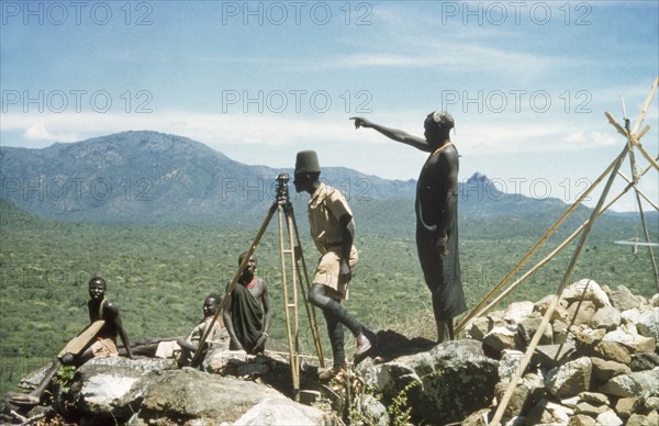 Surveying in the Karasuk hills. Accompanied by Suk (Pokot) porters, a Ugandan Forest Guard stands at the base of a 'kituti' (cairn) marked with bamboo sticks, surveying Sinjalol and Tarakit in the Karasuk hills. Part of a forestry survey team, he uses a theodolite to locate and measure the distance to the next 'kituti'. Kenya, May 1959., East (Uganda), Uganda, Eastern Africa, Africa.