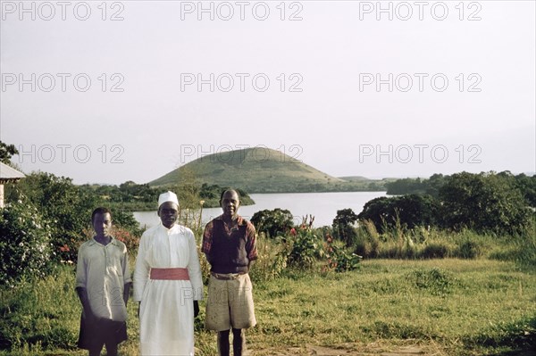 Portrait of three Ugandan house staff. Three Ugandan house staff on the shores of a lake. They are identified (left to right) as: "The toto, who did all the dirty work and cut the grass, ...Asanasio who waited at table and was (a) safari boy, and ...the cook Nikimia". Probably Saka, West Uganda, 1955., West (Uganda), Uganda, Eastern Africa, Africa.