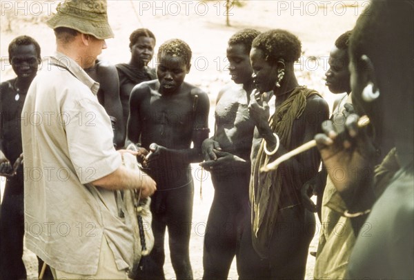 Tobacco exchanged for news. British Game Ranger John Blower offers tobacco to a group of Toposa men at Kabekanyang on the Uganda-Sudan border. In return, he receives the 'habari' (news) about "game movements, poachers, marauders, cattle rustlers". Kabekanyang, North East, Uganda, 1959., North (Uganda), Uganda, Eastern Africa, Africa.