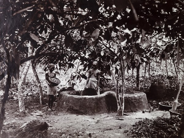 View of two figures with palm kernels by mud enclosure