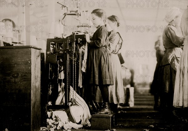 Knitter is a little girl so small she has to stand on a box to reach her machine. 1910