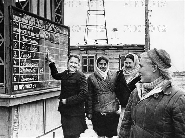 A crew of young women workers, headed by mari volkova (right), have just finished their shift and are reading the results of the high output competition, volkova and her girlfriends are in second place, the heading of the 'exhibition board' reads 'fulfillment quota for oil output from ,,,', 1940s.