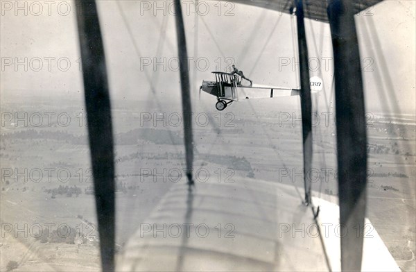 Photograph of Curtiss JN-4 aircraft C187 in flight, with a man [Lieutenant Ned Ballough] 'wing walking': standing with his arms on the top of the wing and his feet behind the back of the cockpit circa 1917.