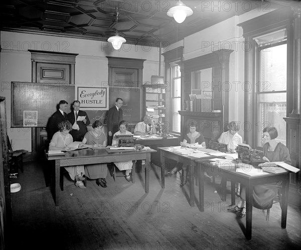 Women typists in an office, "Everybody Hustle" sign in the background ca.  between 1910 and 1935