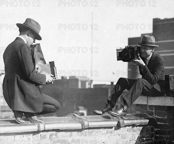 Two photographers taking each other's picture with hand-held cameras while perched on a roof ca. 1909