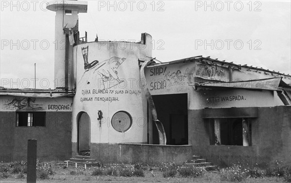 1947 - Ruin of a burnt-out building with graffiti. Boot with nazi cross (swastika) on back of woman with child. Text: Djika Blanda and Djangan Mendjadjah, among others. -  Location: Indonesia, Java, Dutch East Indies