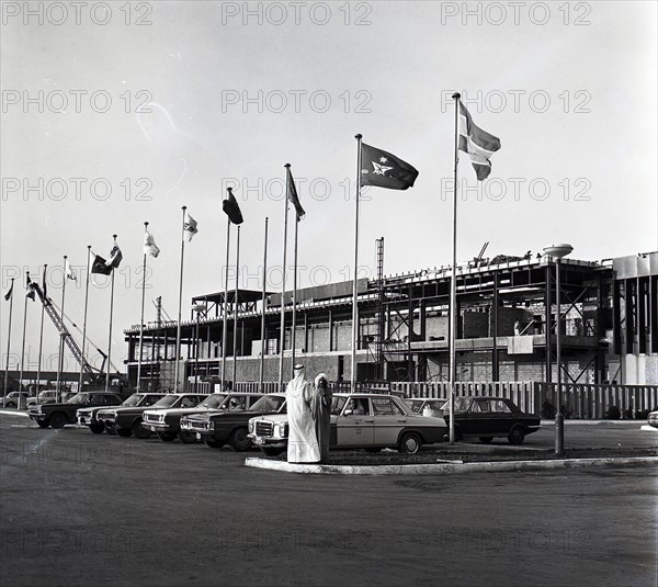 A photograph of the exterior of Bahrain International Airport in December 1975. Parked taxis can be seen in the foreground amongst flag poles