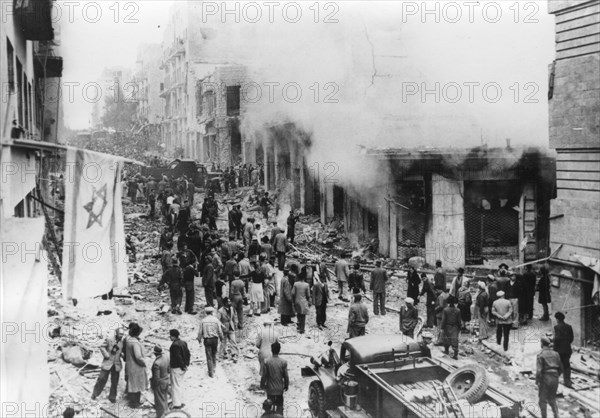 The Jewish Star of David flag hangs from its broken pole across from destroyed buildings and rubble.