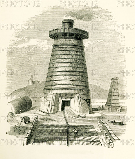 This illustration dates to the 1870s and shows a blast furnace in Great Britain, where the enormous quantity of iron produced from clay ironstone was first obtained in the state of cast iron by the process of smelting. In thisprocess the clay ironstone is roasted after having been broken up into lumps. When ready, the ore is then put in a blast furnace (seen here), a structure about 40 to 50 feet high and 12 to 17 fet in internal diameter at its widest part.