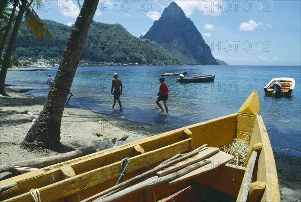WEST INDIES, St Lucia , Soufriere, People walking along beach with boats moored offshore and the Pitons beyond and prow of boat in imediate foreground.